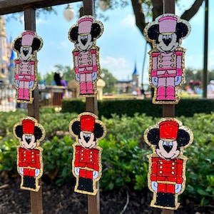 May include: Several Mickey Mouse-shaped ornaments in the image. The ornaments are in the shape of toy soldiers, with red and pink uniforms, black ears, and gold glitter trim. The background shows a park with a castle.