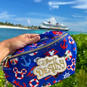 May include: A blue fanny pack featuring a repeating nautical pattern of red and white life preservers, anchors, and Mickey Mouse icons. A gold-trimmed, white textured patch on the front displays the text Disney DESTINY in gold lettering.