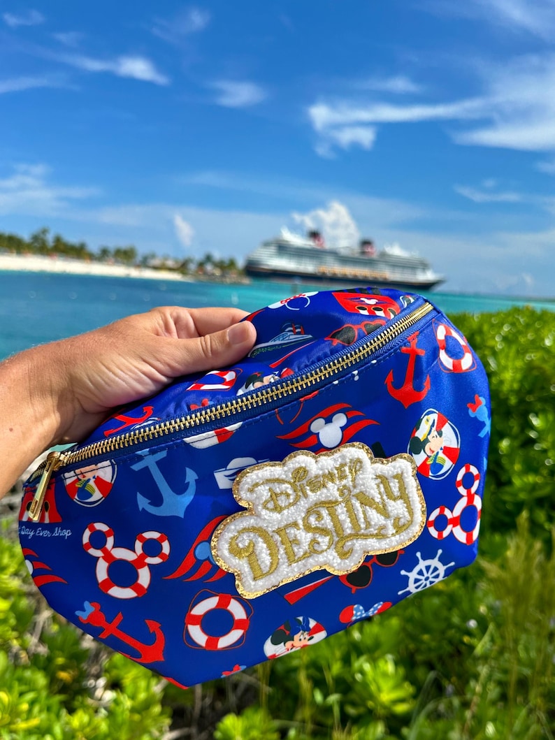May include: A blue fanny pack featuring a repeating nautical pattern of red and white life preservers, anchors, and Mickey Mouse icons. A gold-trimmed, white textured patch on the front displays the text Disney DESTINY in gold lettering.