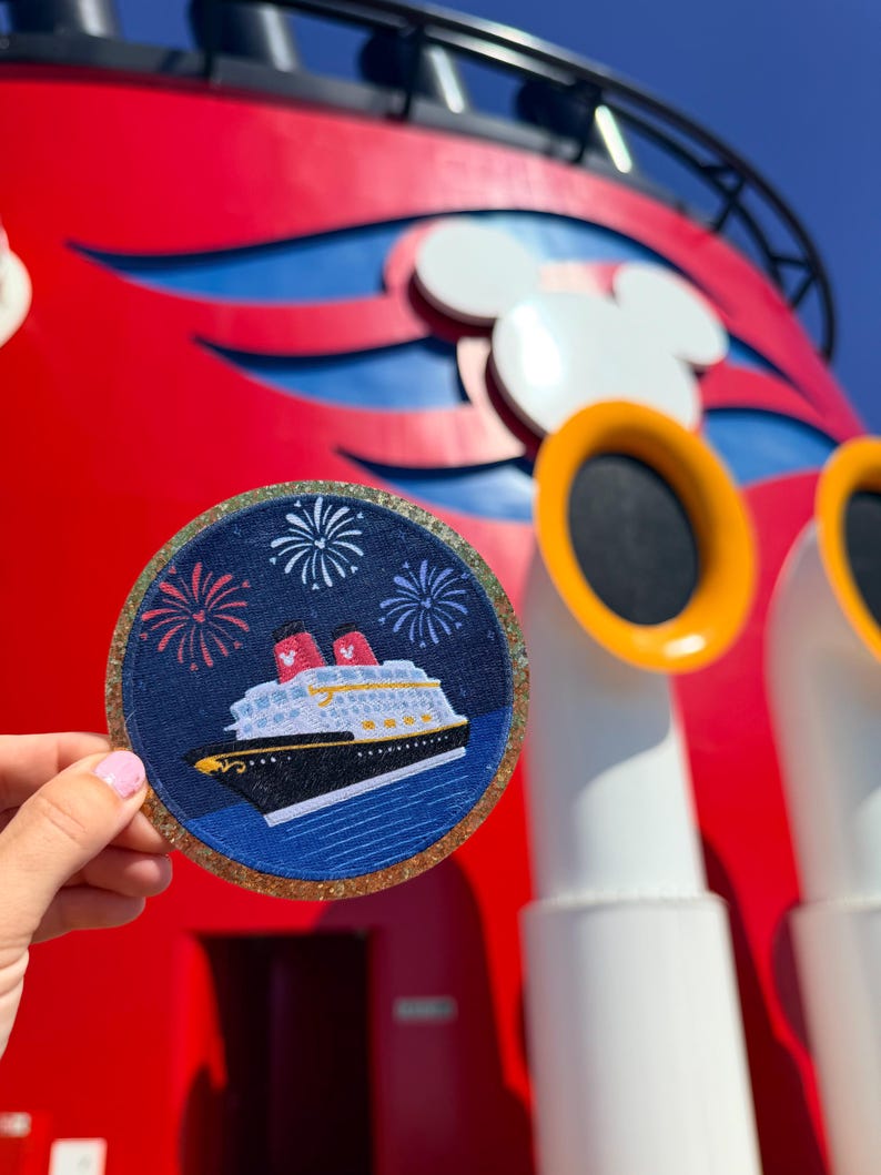 May include: A circular embroidered patch featuring a cruise ship on a dark blue background with white fireworks above. The patch has a gold glitter border and depicts a ship with a black hull, white superstructure, and two red smokestacks.