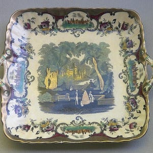 May include: A rectangular ceramic serving tray with a gold rim and handles. The tray features a central landscape scene with figures and a castle, surrounded by floral and architectural border designs in various colors.