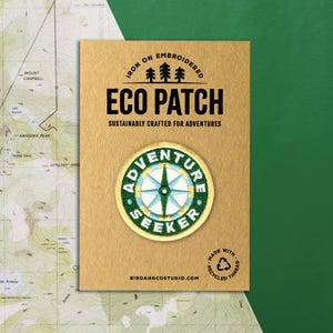 May include: An iron-on embroidered Eco Patch with the text "Adventure Seeker" and a compass design. The patch is on a tan card with the text "Sustainably Crafted for Adventures". The background includes a map and a green surface.