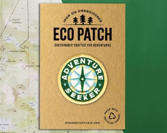 May include: An iron-on embroidered Eco Patch with the text "Adventure Seeker" and a compass design. The patch is on a tan card with the text "Sustainably Crafted for Adventures". The background includes a map and a green surface.