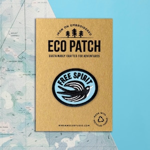 May include: An iron-on embroidered Eco Patch on a tan card. The patch features a black bird design within a light blue oval, with the words "FREE SPIRIT." The card reads "SUSTAINABLY CRAFTED FOR ADVENTURES" and "MADE WITH RECYCLED THREAD."