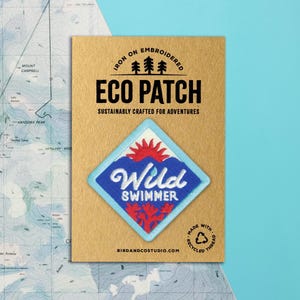 May include: An iron-on embroidered Eco Patch with the text "Wild Swimmer" in white script. The patch features a blue diamond shape with a red sun and coral design. The patch is on a brown card with the text "Sustainably Crafted for Adventures".