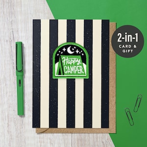 May include: A card with a black and cream striped pattern, featuring a green patch with the words "Happy Camper" and a camping scene. A green pen and two paper clips are also visible.