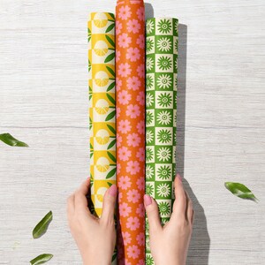 May include: Three rolls of wrapping paper with different patterns. The first roll has a yellow and green pattern with lemons and leaves. The second roll has a pink and orange pattern with flowers. The third roll has a green and white pattern with flowers.