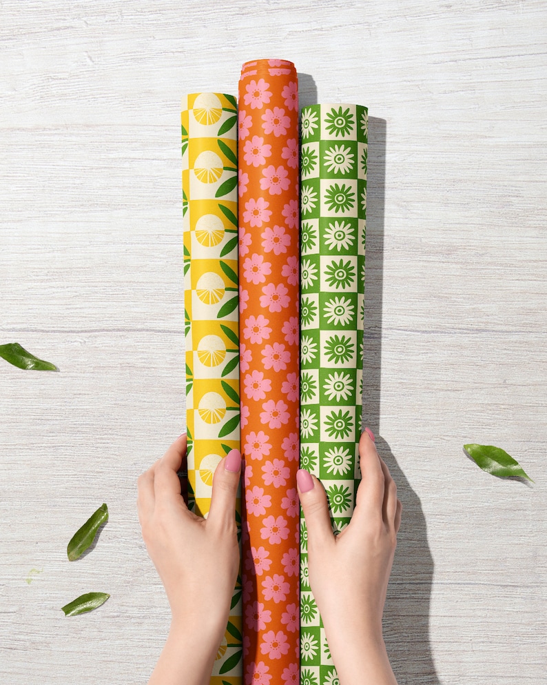 May include: Three rolls of wrapping paper with different patterns. The first roll has a yellow and green pattern with lemons and leaves. The second roll has a pink and orange pattern with flowers. The third roll has a green and white pattern with flowers.