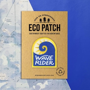 May include: An embroidered Eco Patch with a blue and yellow wave design, the words "WAVE RIDER" are displayed. The patch is on a brown card with the text "SUSTAINABLY CRAFTED FOR ADVENTURES". The background is a blue surface and a map.