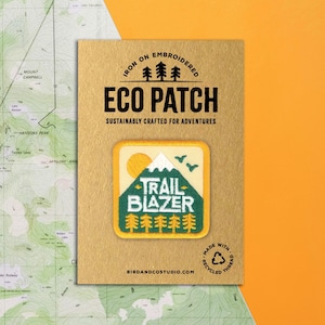 May include: An embroidered Eco Patch with the words "TRAIL BLAZER" on a square patch with a mountain and tree design. The patch is on a brown card with the text "SUSTAINABLY CRAFTED FOR ADVENTURES".