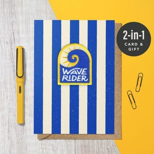 May include: A greeting card with blue and white stripes, featuring a patch with a wave design and the words "WAVE RIDER." A yellow pen and two paper clips are also present. The card is labeled "2-in-1 CARD & GIFT."