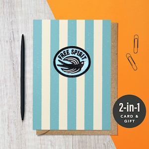 May include: A greeting card with blue and cream vertical stripes, featuring a circular emblem with a bird and the words "FREE SPIRIT." A black pen and paper clips are on an orange background. The card is labeled "2-in-1 CARD & GIFT."
