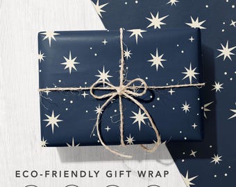 May include: A rectangular gift wrapped in dark navy blue paper featuring a pattern of cream-colored stars and dots, tied with natural jute twine. Below, the text reads ECO-FRIENDLY GIFT WRAP with four circular icons for recycled, carbon balanced, biodegradable, and sustainable paper.