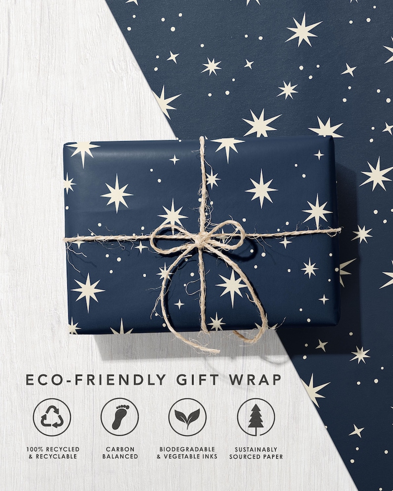 May include: A rectangular gift wrapped in dark navy blue paper featuring a pattern of cream-colored stars and dots, tied with natural jute twine. Below, the text reads ECO-FRIENDLY GIFT WRAP with four circular icons for recycled, carbon balanced, biodegradable, and sustainable paper.