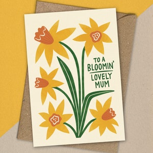 Puede incluir: Una tarjeta con un dise&ntilde;o floral con narcisos amarillos con centros naranjas y tallos verdes. El texto "To a Bloomin' Lovely Mum" est&aacute; impreso en la tarjeta.