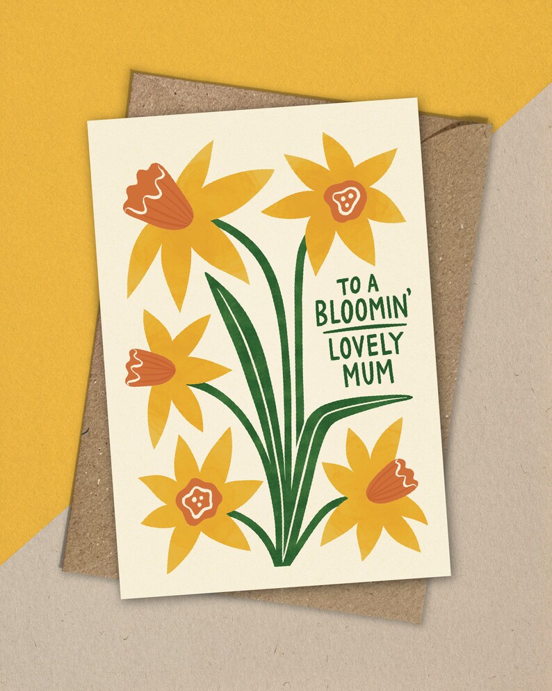 Puede incluir: Una tarjeta con un dise&ntilde;o floral con narcisos amarillos con centros naranjas y tallos verdes. El texto "To a Bloomin' Lovely Mum" est&aacute; impreso en la tarjeta.