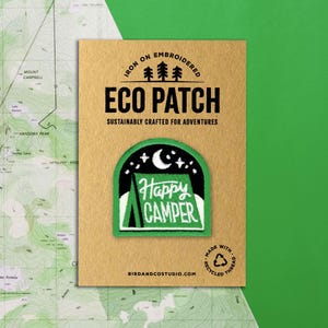 May include: An embroidered "Happy Camper" eco patch on a tan card. The patch shows a green tent under a night sky with a crescent moon and stars. The card reads "Iron On Embroidered Eco Patch" and "Sustainably Crafted for Adventures."