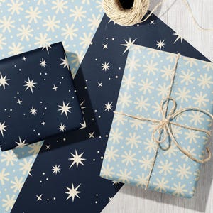 May include: Two rectangular gift boxes wrapped in paper with star and snowflake patterns. One box features a dark navy background with cream-colored stars, while the other is light blue with cream snowflakes, tied with natural jute twine.