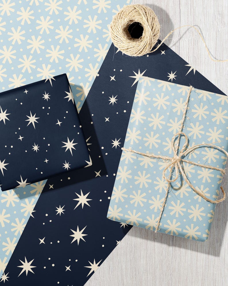 May include: Two rectangular gift boxes wrapped in paper with star and snowflake patterns. One box features a dark navy background with cream-colored stars, while the other is light blue with cream snowflakes, tied with natural jute twine.