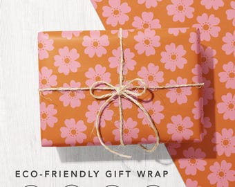 May include: A rectangular gift wrapped in orange paper with a repeating pink flower pattern, tied with natural twine. Below, text reads ECO-FRIENDLY GIFT WRAP with four circular icons representing 100% recycled, carbon balanced, biodegradable, and sustainably sourced paper.