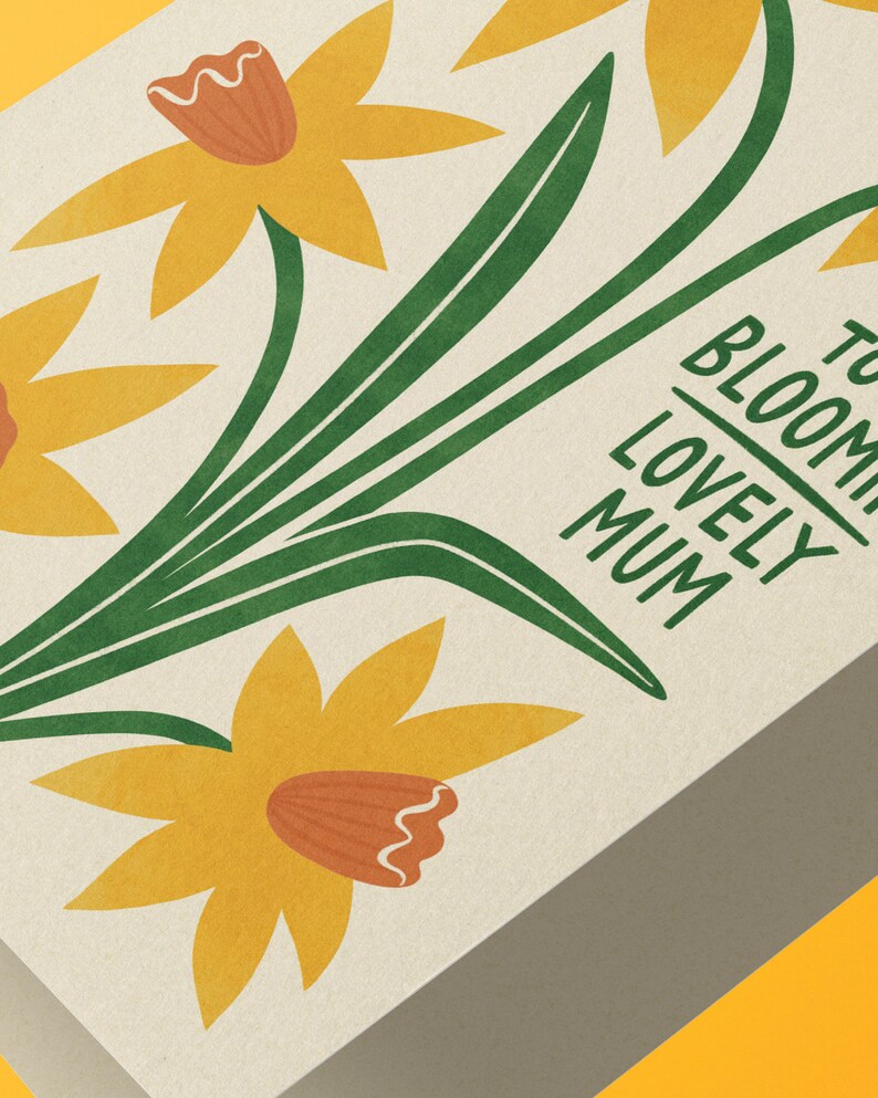 Puede incluir: Una tarjeta con un dise&ntilde;o floral con narcisos amarillos con centros naranjas y tallos verdes. El texto "To Bloom Lovely Mum" est&aacute; impreso en la tarjeta.
