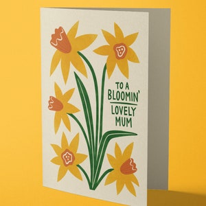 Puede incluir: Una tarjeta con un dise&ntilde;o floral con narcisos amarillos con centros naranjas y tallos verdes. El texto de la tarjeta dice "To a Bloomin' Lovely Mum".