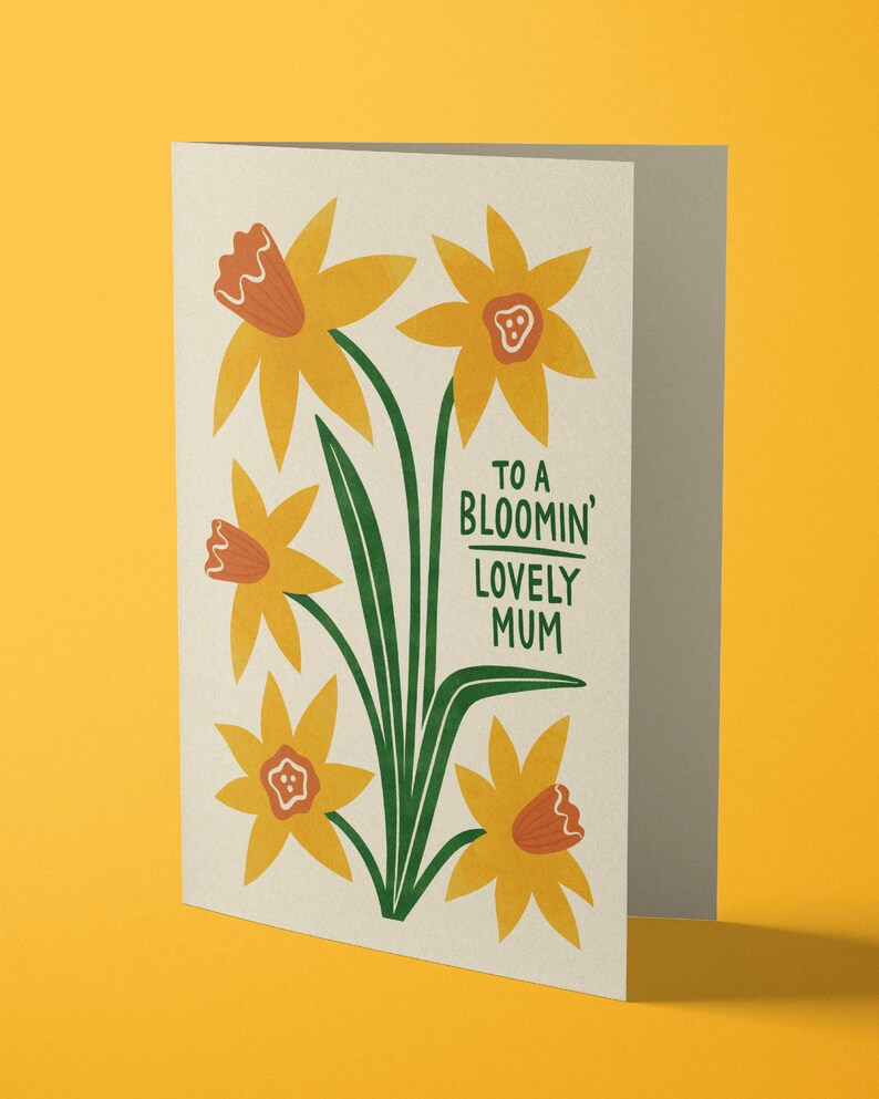 Puede incluir: Una tarjeta con un dise&ntilde;o floral con narcisos amarillos con centros naranjas y tallos verdes. El texto de la tarjeta dice "To a Bloomin' Lovely Mum".
