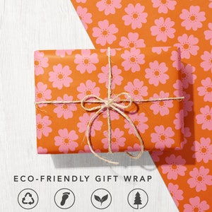 May include: A gift wrapped in orange paper with a pink floral pattern. The paper is tied with twine and the gift is sitting on a white wooden surface. The text "ECO-FRIENDLY GIFT WRAP" is below the gift. There are four icons below the text, each with a different symbol and text: 100% RECYCLED & RECYCLABLE, CARBON BALANCED, BIODEGRADABLE & VEGETABLE INKS, FSC&reg; CERTIFIED PAPER.