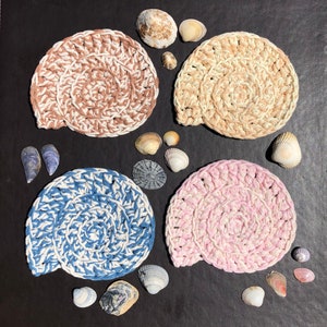 May include: Four crocheted coasters in different colors and patterns. The coasters are round and have a spiral design. They are arranged in a square pattern on a black surface. There are also several seashells scattered around the coasters.