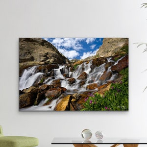 May include: A framed landscape photograph of a cascading waterfall over rocks, with a blue sky and clouds in the background. The image is displayed in a modern living room setting with a green chair, glass coffee table, and a large plant.