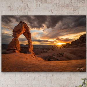 May include: A canvas print of a desert landscape at sunset. The image features a natural sandstone arch under a dramatic sky with the sun setting in the distance. The print is displayed above a wooden bench with woven baskets.