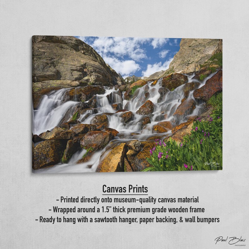 May include: Canvas print of a cascading waterfall flowing over rocks, with a blue sky and clouds in the background. The print is wrapped around a 1.5-inch thick wooden frame. The image includes the text "Canvas Prints" and the artist's signature.