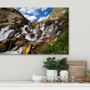 May include: A canvas print of a mountain waterfall cascading over rocks. The water is blurred, creating a silky effect. The rocks are various shades of brown and gold, with green vegetation and purple flowers. A blue sky with clouds is visible.