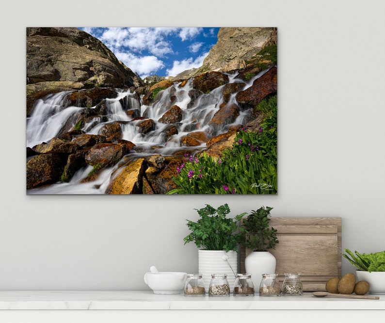 May include: A canvas print of a mountain waterfall cascading over rocks. The water is blurred, creating a silky effect. The rocks are various shades of brown and gold, with green vegetation and purple flowers. A blue sky with clouds is visible.