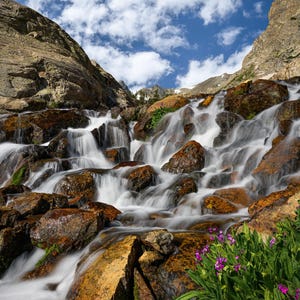 May include: A scenic landscape photograph of a cascading waterfall flowing over large, brown rocks. The water is a blurred white, contrasting with the rocks. Green plants and purple flowers are in the foreground, with a blue sky and clouds above.