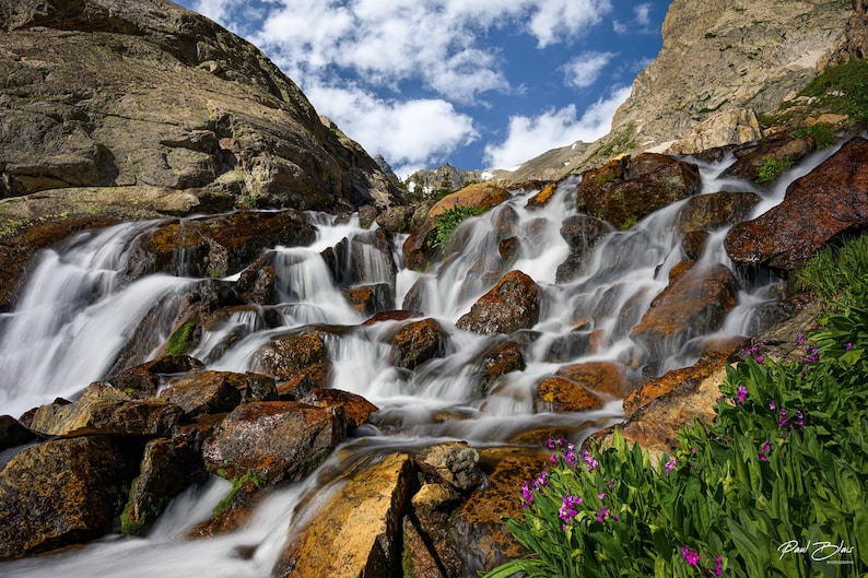 May include: A scenic landscape photograph of a cascading waterfall flowing over large, brown rocks. The water is a blurred white, contrasting with the rocks. Green plants and purple flowers are in the foreground, with a blue sky and clouds above.