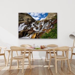 May include: A framed canvas print of a waterfall cascading over rocks, with a blue sky and clouds visible. The print is displayed above a wooden dining table with chairs. The room has hardwood floors and potted plants.