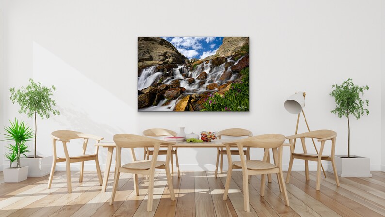 May include: A framed canvas print of a waterfall cascading over rocks, with a blue sky and clouds visible. The print is displayed above a wooden dining table with chairs. The room has hardwood floors and potted plants.