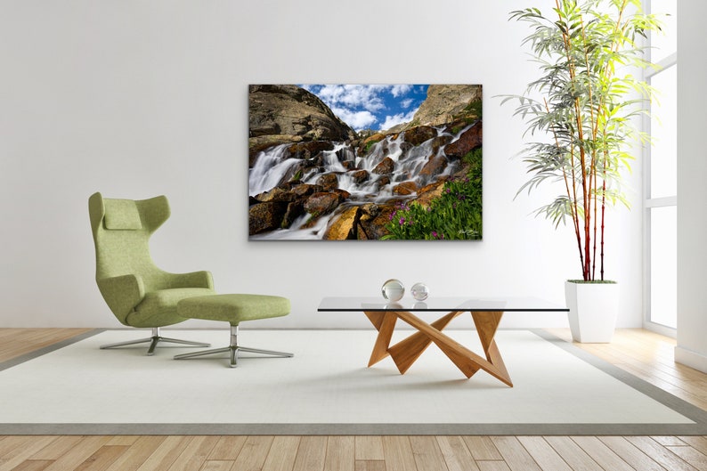 May include: A framed landscape photograph of a cascading waterfall over rocks, with a blue sky and clouds in the background. The image is displayed in a modern living room setting with a green chair, glass coffee table, and a large plant.