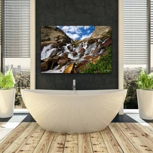 May include: A modern bathroom with a large, white, oval bathtub and a framed landscape photograph of a waterfall. The room has a wooden floor, large windows with blinds, and two potted plants.