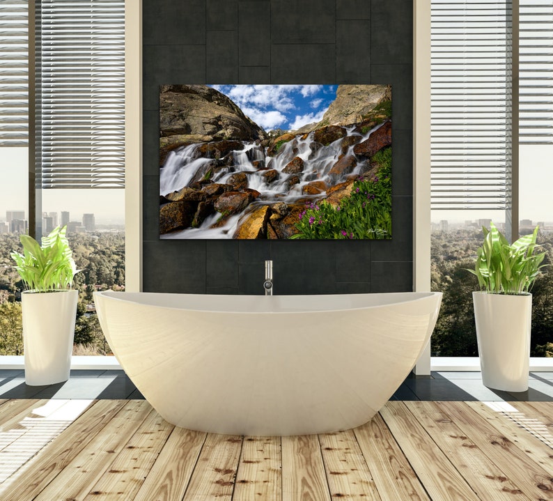 May include: A modern bathroom with a large, white, oval bathtub and a framed landscape photograph of a waterfall. The room has a wooden floor, large windows with blinds, and two potted plants.