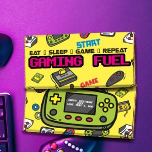 Op de afbeelding: Een gele en groene chocoladebar-verpakking met een retro videogame-thema. De verpakking heeft een pixelachtig ontwerp met de tekst "START EAT SLEEP GAME REPEAT GAMING FUEL". De verpakking bevat ook een kleiner beeld van een videogamecontroller met de tekst "HAPPY BIRTHDAY JACK LOVE MUM & DAD".