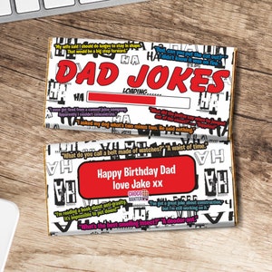 Op de afbeelding: Een witte chocolade reep met een rood en zwart ontwerp. De reep heeft de tekst "Dad Jokes" erop gedrukt. De reep heeft ook een voortgangsbalk met de tekst "Loading..." en verschillende vadersmoppen erop gedrukt. De reep is verpakt in wit papier met een rood en zwart ontwerp. De reep is een cadeau voor een vader en heeft de tekst "Happy Birthday Dad love Jake xx".
