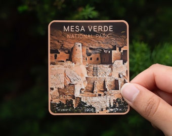 May include: A sticker featuring a colorful illustration of Mesa Verde National Park, with the text "Mesa Verde National Park" and coordinates "37.2309° N 108.4818° W".