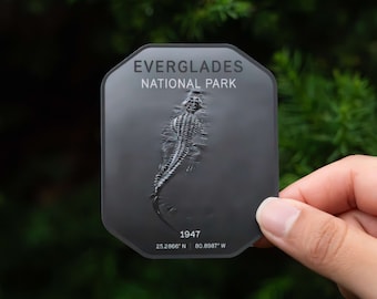May include: A black and white sticker with a grey background featuring an alligator silhouette and the text "EVERGLADES NATIONAL PARK" and "1947 25.2866° N 80.8987° W".