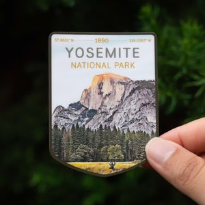 May include: A rectangular sticker featuring an illustration of Yosemite National Park. The image shows a mountain, trees, and a deer in a field. The sticker has the words "YOSEMITE NATIONAL PARK" and coordinates.