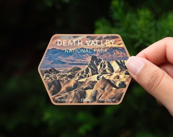 May include: A hexagonal sticker with a scenic image of Death Valley National Park. The sticker features a sunset over a mountain range with a brown, orange, and blue color scheme. The text "Death Valley National Park" is printed in white on the sticker. The coordinates "36.5322° N 116.9325° W" and the year "1994" are printed in white at the bottom of the sticker.