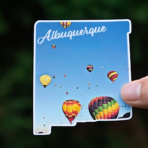 May include: A sticker featuring a blue sky with hot air balloons in various colors and patterns. The sticker is shaped like the state of New Mexico and has the word "Albuquerque" written in white cursive at the top.