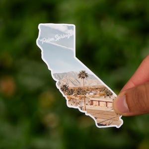 May include: A sticker in the shape of California, featuring a landscape of Palm Springs. The sticker shows a palm tree, mountains, and buildings under a blue sky. The words "Palm Springs" are written across the top.