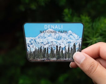 May include: A sticker featuring a scenic view of Denali National Park in Alaska. The sticker shows a snow-capped mountain range with a blue sky and green trees in the foreground. The text "Denali National Park" is printed in black at the top of the sticker, and the coordinates "63.1148°N 151.1928°W" are printed at the bottom.